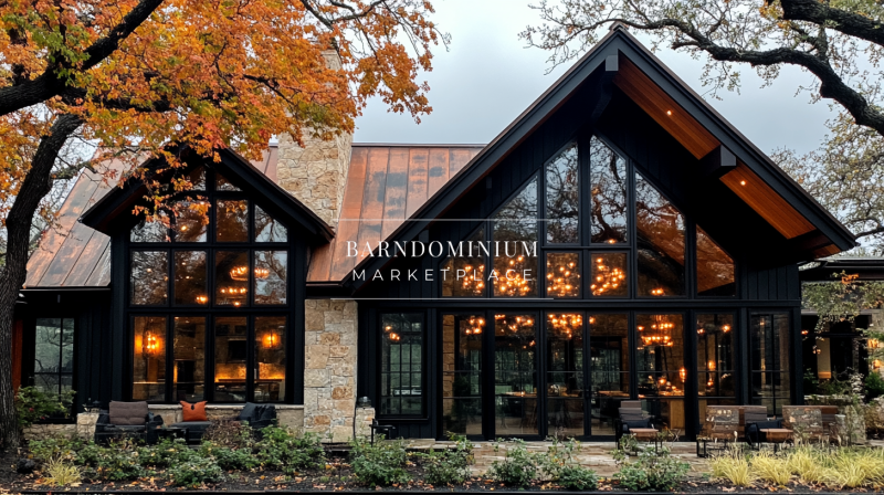A stunning modern black barndominium with large glass windows, a rustic stone chimney, and warm chandelier lighting, set against an autumn backdrop.