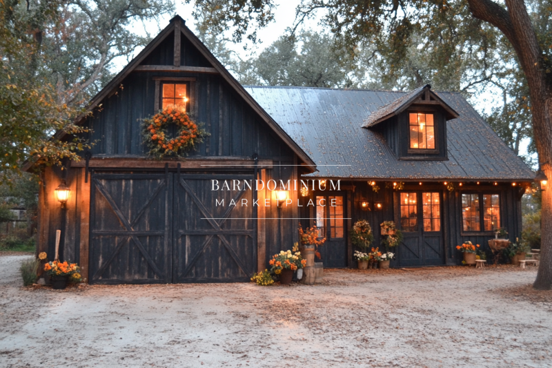 A charming black barndominium with rustic wooden barn doors, warm exterior lighting, and festive fall decor, nestled under large oak trees.