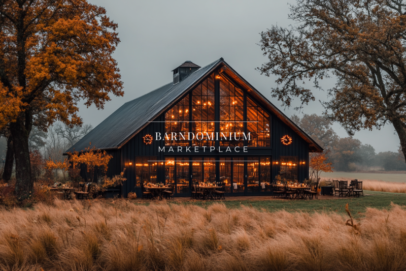 A stunning modern black barn with expansive glass windows, warmly lit from within, set against a backdrop of golden autumn fields and trees.
