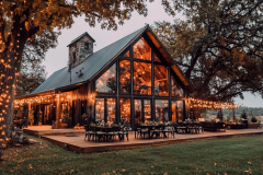 A beautifully lit A-frame barndominium with expansive glass walls, an inviting wraparound deck, and magical string lights under a twilight sky.