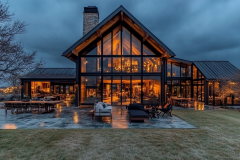 A luxurious modern glass barndominium with warm interior lighting, sleek black metal framing, and a spacious outdoor living area.