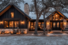 A stunning black modern farmhouse barndominium with warm lighting, stone accents, and a cozy winter setting.