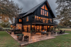 Stunning black modern barndominium with large windows, warm interior lighting, and a spacious outdoor patio for entertaining.