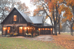 A charming rustic barndominium with a black metal roof, warm glowing lights, and a cozy autumn setting, surrounded by vibrant fall foliage.