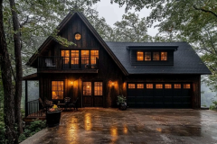 A modern rustic black barndominium with a warm, inviting glow, nestled in a wooded setting on a rainy evening, featuring a garage and upper balcony.