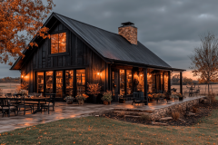 A modern black barndominium with warm lighting, large windows, and a cozy rustic charm, set in a peaceful countryside landscape under an overcast sky.