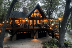 A stunning barndominium-style cabin with a wraparound deck, glowing string lights, and a cozy ambiance nestled among tall trees.