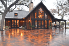 A charming modern farmhouse with a rustic wood exterior, large glass windows, and a cozy, illuminated interior, set against a serene landscape.