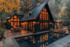 A modern black barndominium nestled in the woods, featuring a sleek metal roof, warm glowing windows, and a luxurious pool reflecting the autumn foliage.