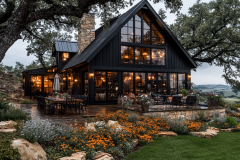 Modern black barndominium with floor-to-ceiling windows, warm lighting, and lush landscaping, blending rustic charm with contemporary elegance.