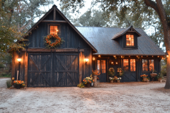 A charming black barndominium with rustic wooden barn doors, warm exterior lighting, and festive fall decor, nestled under large oak trees.