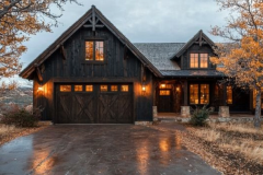 A stunning dark wood barndominium with warm glowing lights, a spacious garage, and a cozy rustic charm, set against an autumn landscape.