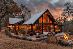 Modern rustic cabin with floor-to-ceiling windows, warm lighting, and an outdoor fire pit, set against a dramatic sunset sky.