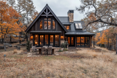 A charming black barndominium nestled in an autumn landscape, featuring large windows, warm lighting, and a cozy outdoor dining area.