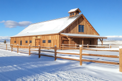 A stunning wooden barndominium exterior covered in snow, featuring a classic barn-style design with a rustic touch.