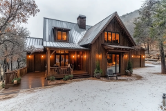 A rustic winter barndominium with warm glowing lights, wooden siding, a metal roof, and a cozy front porch surrounded by a snowy landscape.