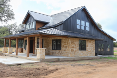 A beautifully designed barndominium with a mix of natural stone and black board-and-batten siding, complemented by a sleek metal roof and a spacious covered porch.