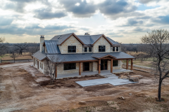 A charming two-story barndominium with a rustic stone facade, black metal roof, and warm wooden accents, set against a peaceful countryside backdrop.