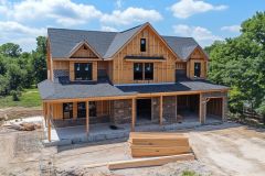 A two-story barndominium in the early stages of construction, featuring a wraparound porch and rustic wooden framing.