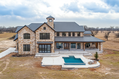 Luxurious barndominium with stone exterior, black metal roof, and a backyard pool, blending rustic charm with modern elegance.