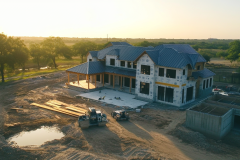 Barndominium under construction at sunset, featuring a stone exterior, wraparound porch, and modern farmhouse design.
