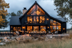 Modern black barndominium with floor-to-ceiling windows, warm ambient lighting, and a stunning outdoor dining space.
