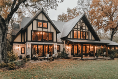 Elegant Tudor-style barndominium with warm lighting, large windows, and a cozy autumn landscape.