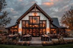 Stunning modern barndominium with large glass windows, warm ambient lighting, and a beautifully designed outdoor dining area.