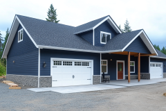 Charming navy blue barndominium with white garage doors and a covered porch, nestled in a wooded setting.