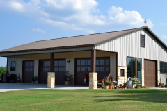 Spacious beige and brown barndominium with three garage doors, a covered porch, and a well-maintained lawn, combining functionality with modern rural living.