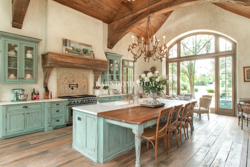 A stunning French country-inspired barndominium kitchen featuring soft mint-green cabinetry, a grand wood range hood, rustic wood beams, and elegant chandeliers.