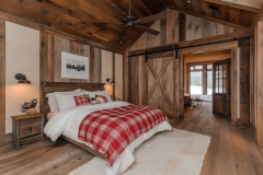 A cozy rustic barndominium bedroom with warm wood paneling, a barn door entrance, and a red plaid bedspread for a charming farmhouse touch.