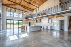 A bright and spacious barndominium interior featuring polished concrete floors, vaulted ceilings, a modern open kitchen, and a mezzanine with black railing.