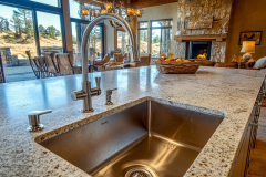 A spacious barndominium kitchen featuring a granite island with a stainless steel sink, seamlessly flowing into a cozy living area with a stone fireplace.