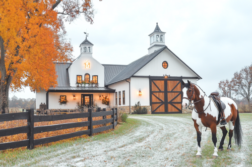 A stunning white barndominium with black trim, warm glowing lights, and a classic barn-style garage, featuring a fenced pasture and a saddled horse in the foreground.