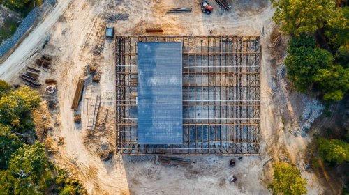An aerial view of a barndominium under construction, showcasing a partially completed steel frame structure with construction materials and equipment scattered around the site.